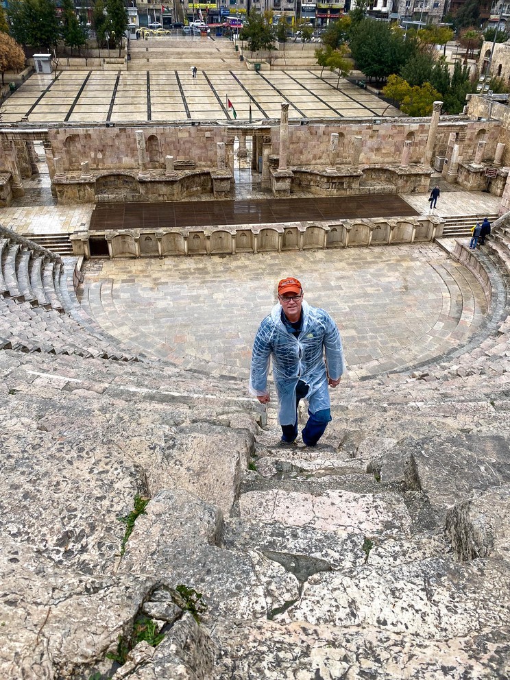 a man tries to climb the limbstone stairs of an ancient Roman theater without breaking the cheap white plastic pancho he is wearing