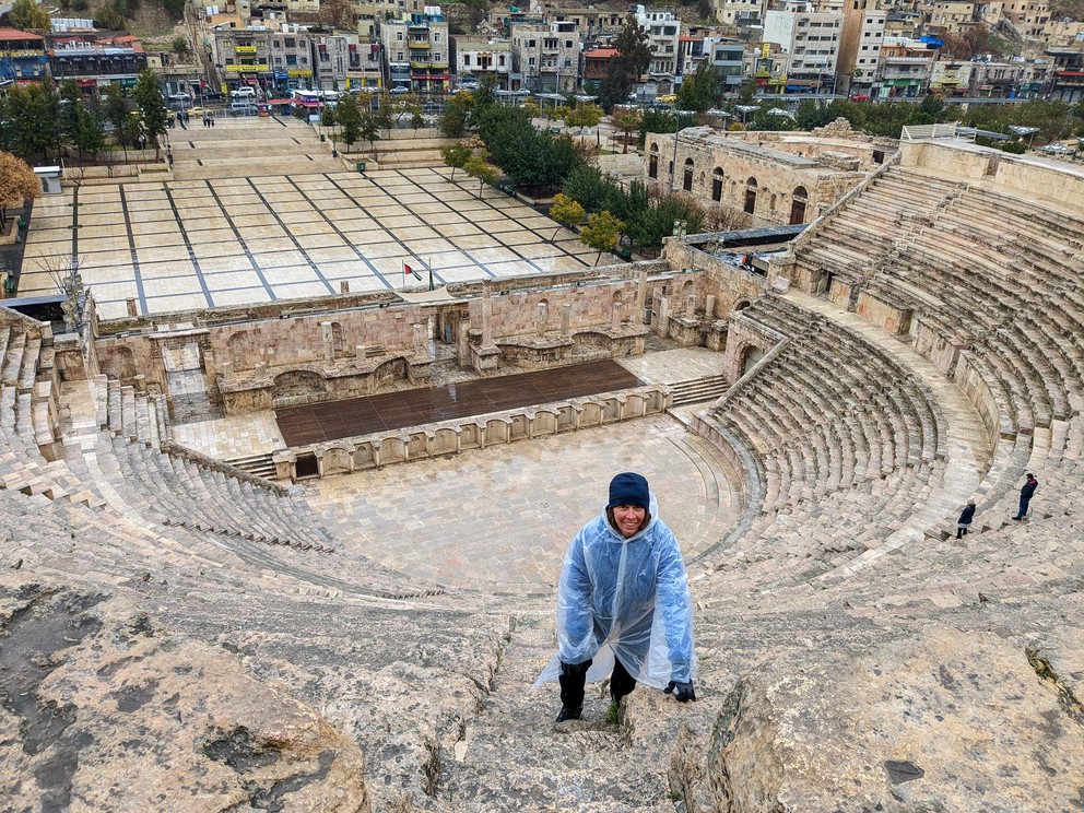 a woman in a white plastic pancho smiles as she climbs the limestone stairs in an ancient Roman Theater
