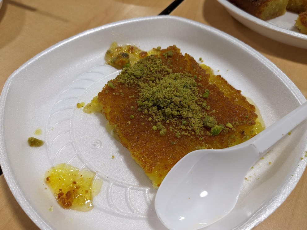 a white styrofoam plate with a healthy square of Kunafa with honey dripping out and some pistachio dust on top