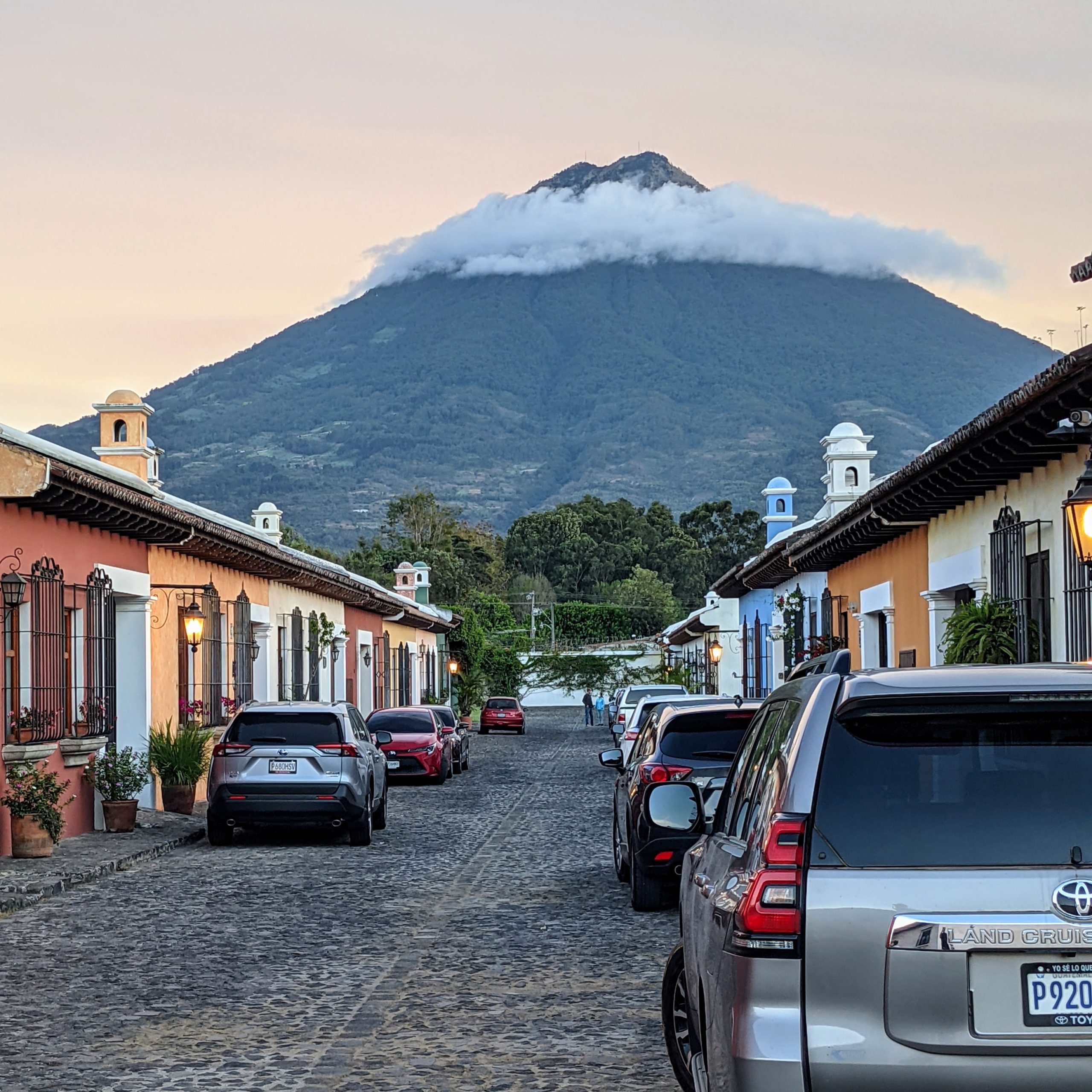 a warm sky at sunrise surrounds a volcano rising above a wide cobblestone street lined with nice cars and colorful houses in Antigua, Guatemala