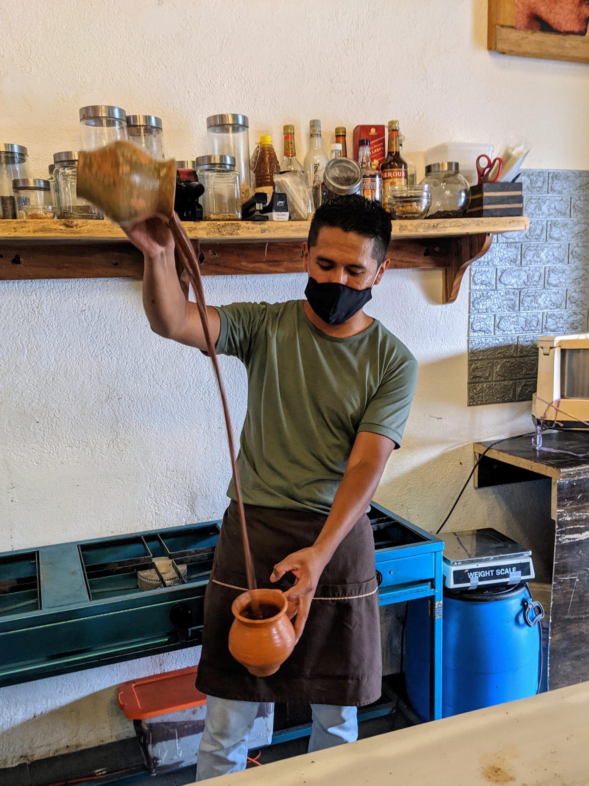 a chocolatier pours liquid chocolate from a pot high in the air into another pot below his waist