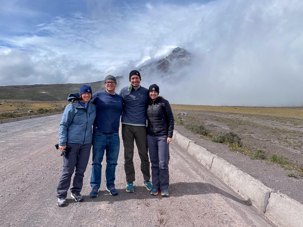 four people stand on a gravel road with a volcano obscured by clouds behind them
