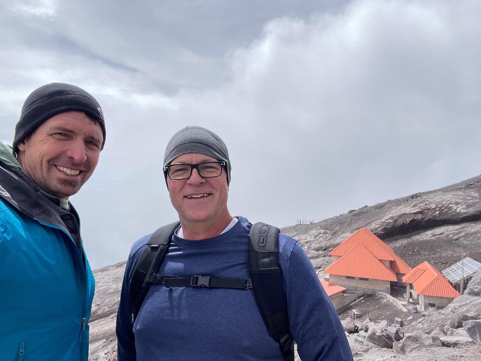 two men stand above a lone building high on the side of a volcano