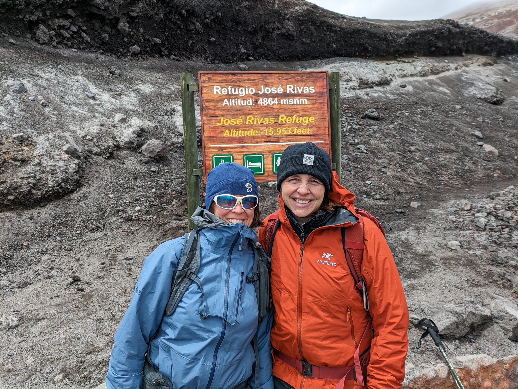 two people stand in volcanic ash in front of a sign indicating a climbers' refuge high on the mountainside