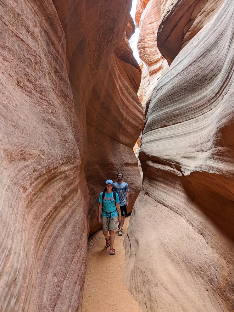 a woman and man walk through a narrow sandy bottom slot canyon