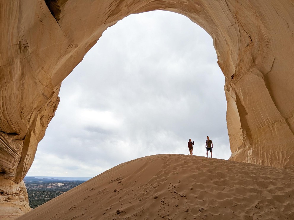 two people stand on a pile of sand inside a large cavern made of yellow sandstone