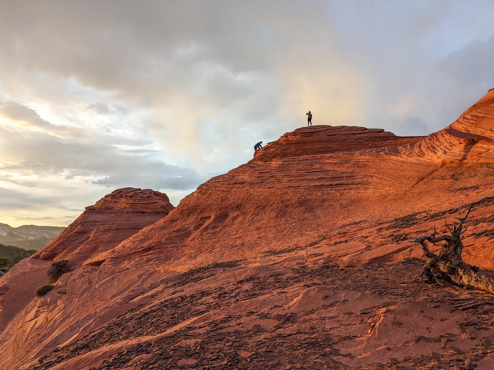 two people ascend a large orange sandstone bluff as the sun sets