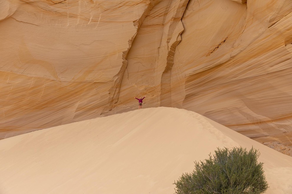 a woman raises her arms in celebration as she stands atop a large pile of buttery yellow sand with a huge yellow sandstone wall behind her