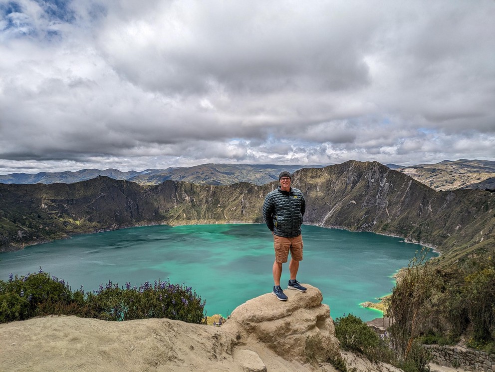 a hiker stands on a rock outcropping high above a deep emerald green volcanic lake