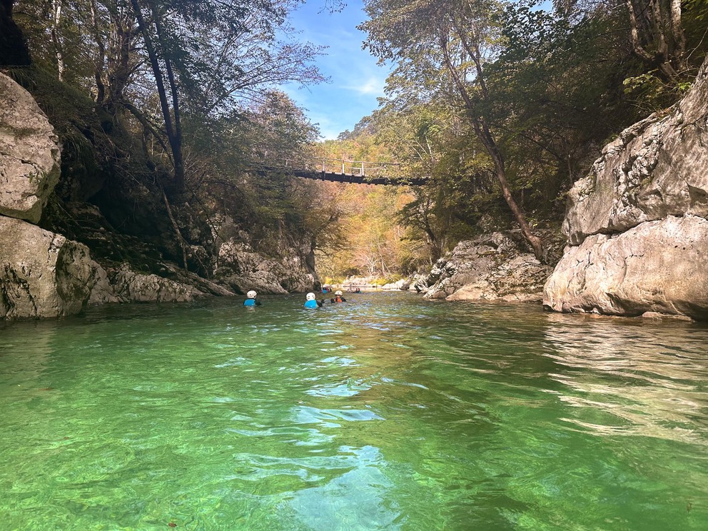 a group of adventurers float on an emerald green river as it passes under a suspension bridge
