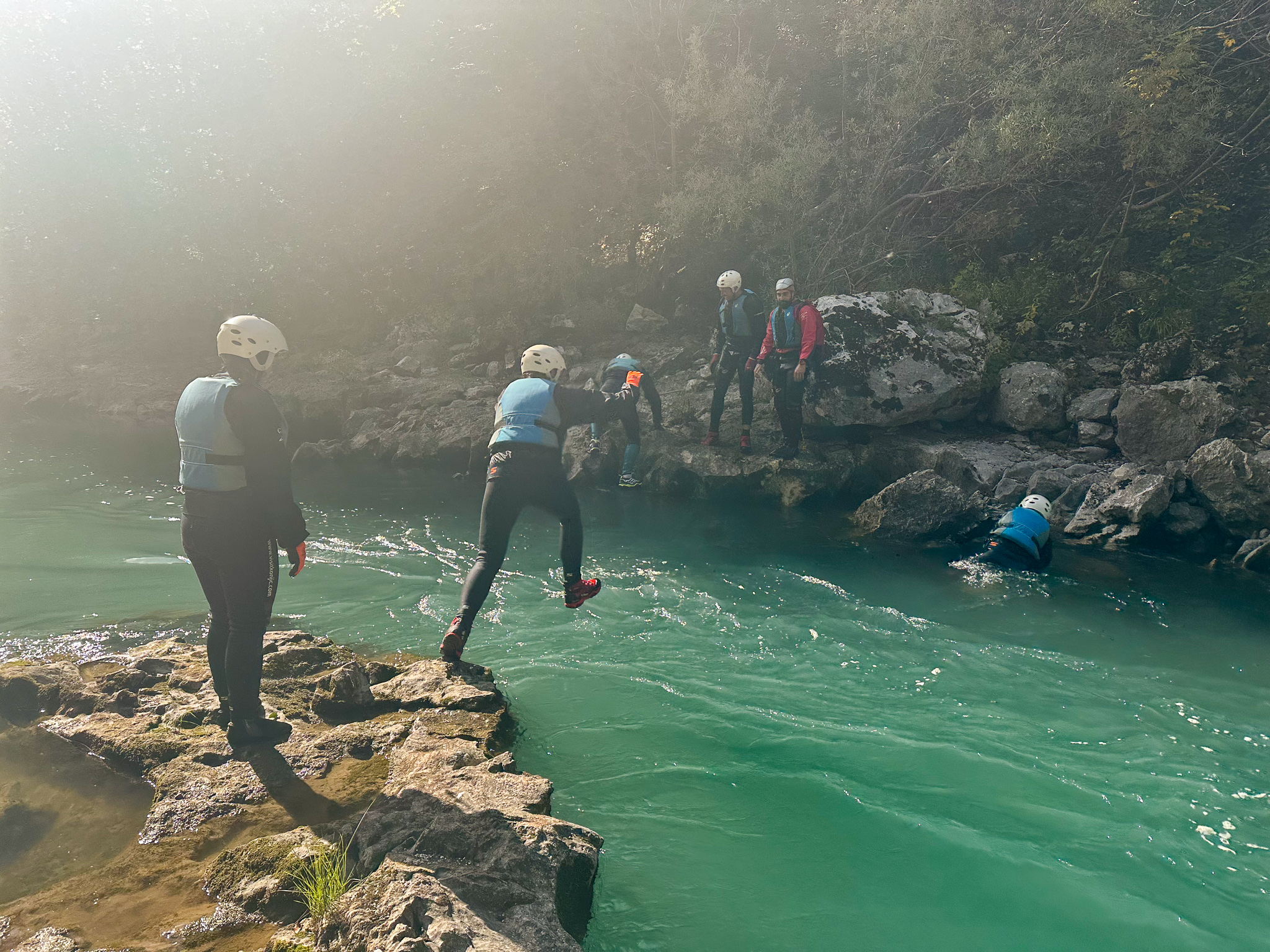 several adventurers wearing wetsuits, life jackets, helmets, and gloves jump into and cross a beautiful emerald green river