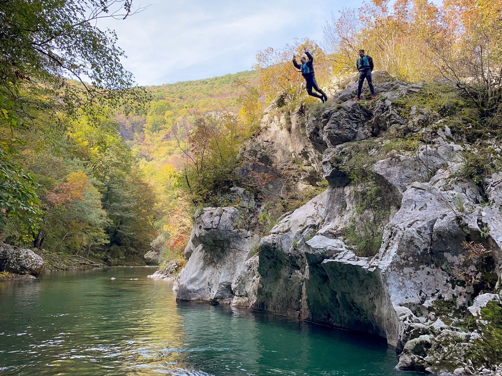 a man jumps from a rocky outcropping about 8 meters above a beautiful turquoise river in the canyon below