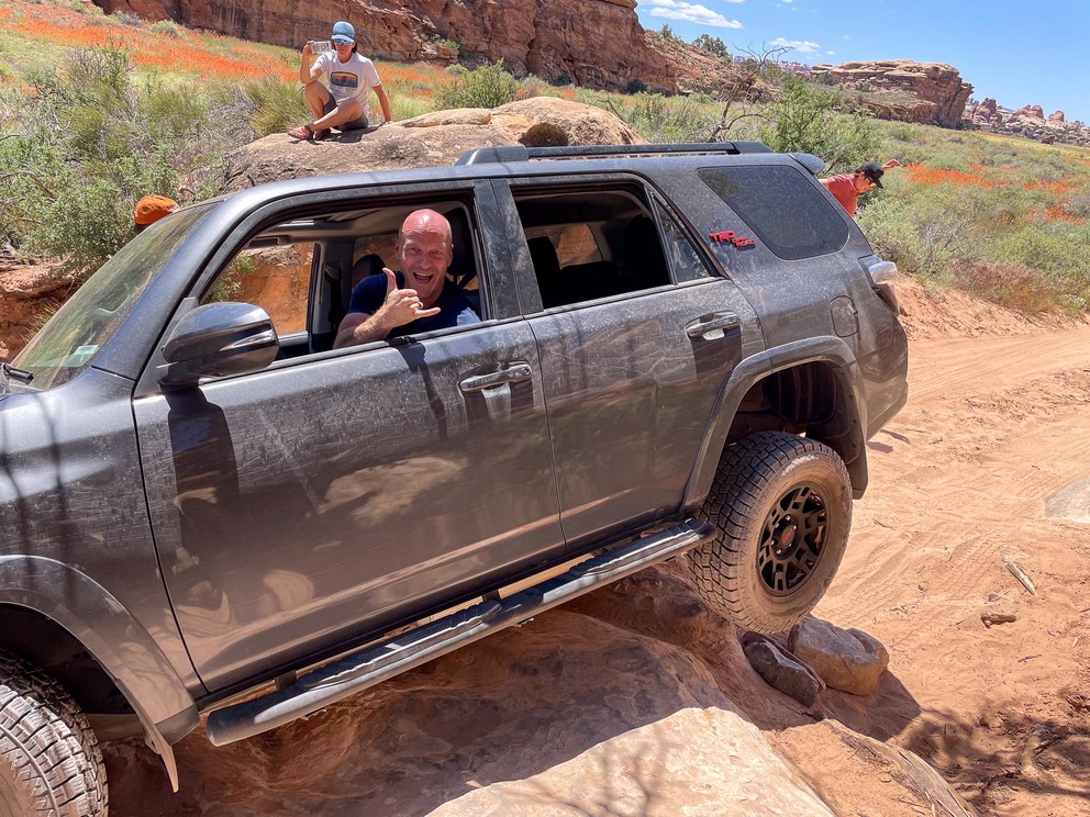 the driver of a Toyota 4Runner gives the Shaka sign with his rear tire suspended in the air as he clears a large obstacle