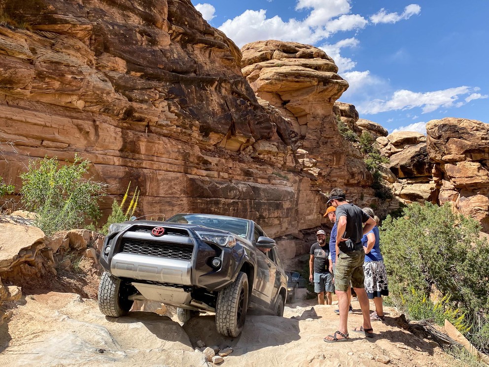 several people watch as a stock Toyota 4Runner attempts to climb over some large rocks next to a sandstone wall