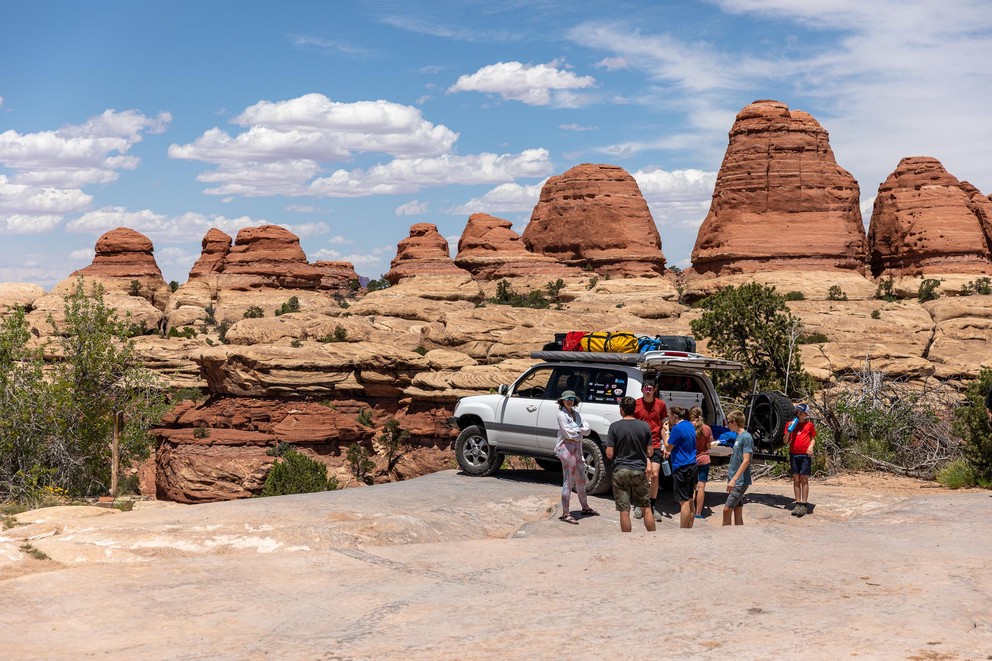 a group of people stand next to a Toyota Land Cruiser with red and yellow sandstone formations behind them