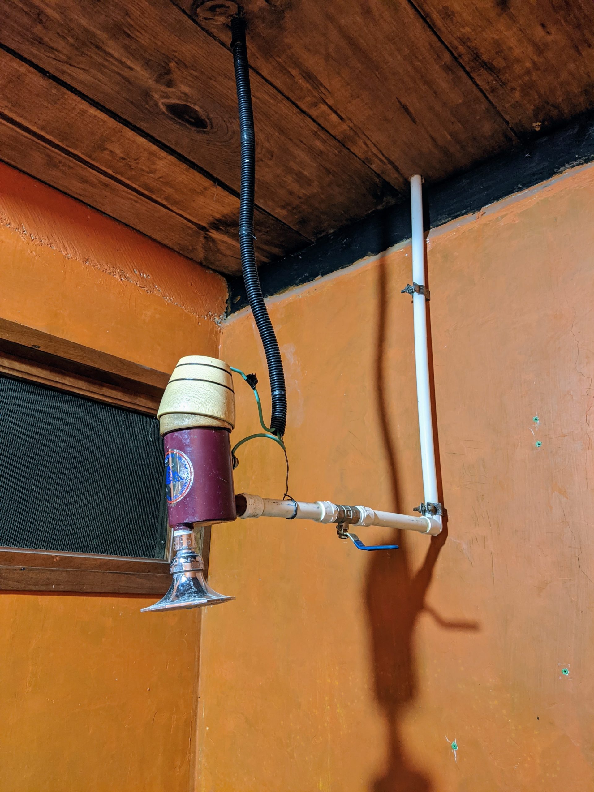 electrical wires come down from the ceiling and attach to a showerhead at the end of a PVC pipe extending out from the wall