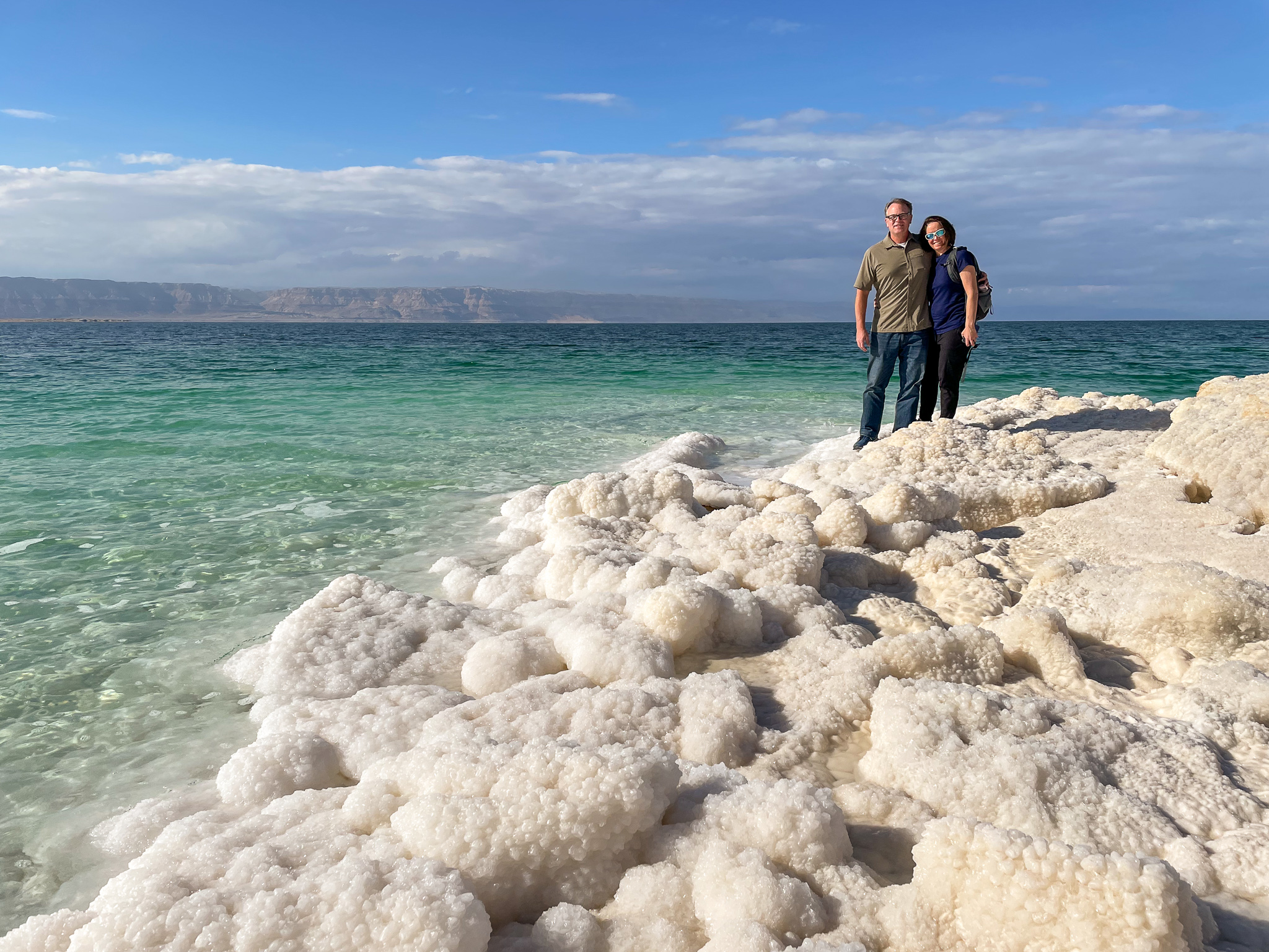 a man and woman stand on a beach made entirely of salt with the turquoise waters of the Dead Sea behind them