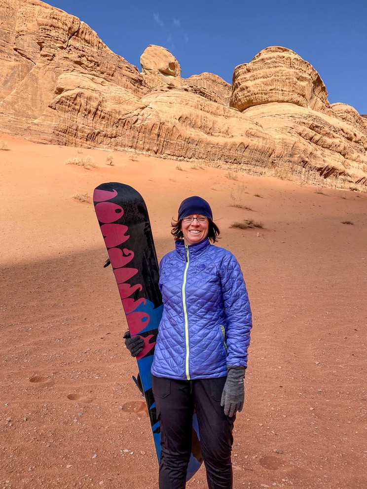 a woman holding a snowboard wears a coat, hat, and gloves while standing near a steep orange hill of sand in the desert