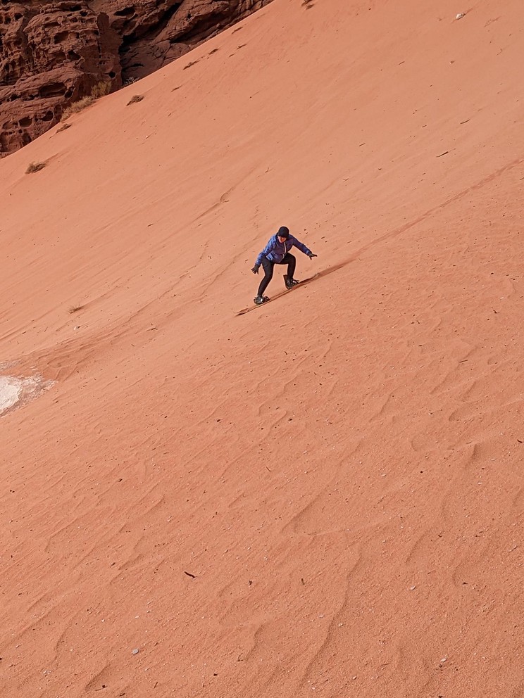 a woman rides a snowboard down a steep orange sand hill in the desert