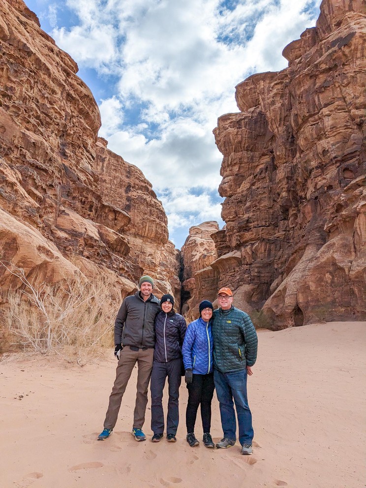 four tourists dressed in coats and hats stand in the sand in front of a narrow canyon