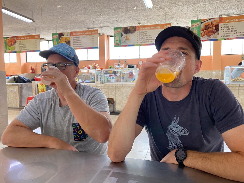 two men drink orange liquid from plastic cups inside a cafeteria