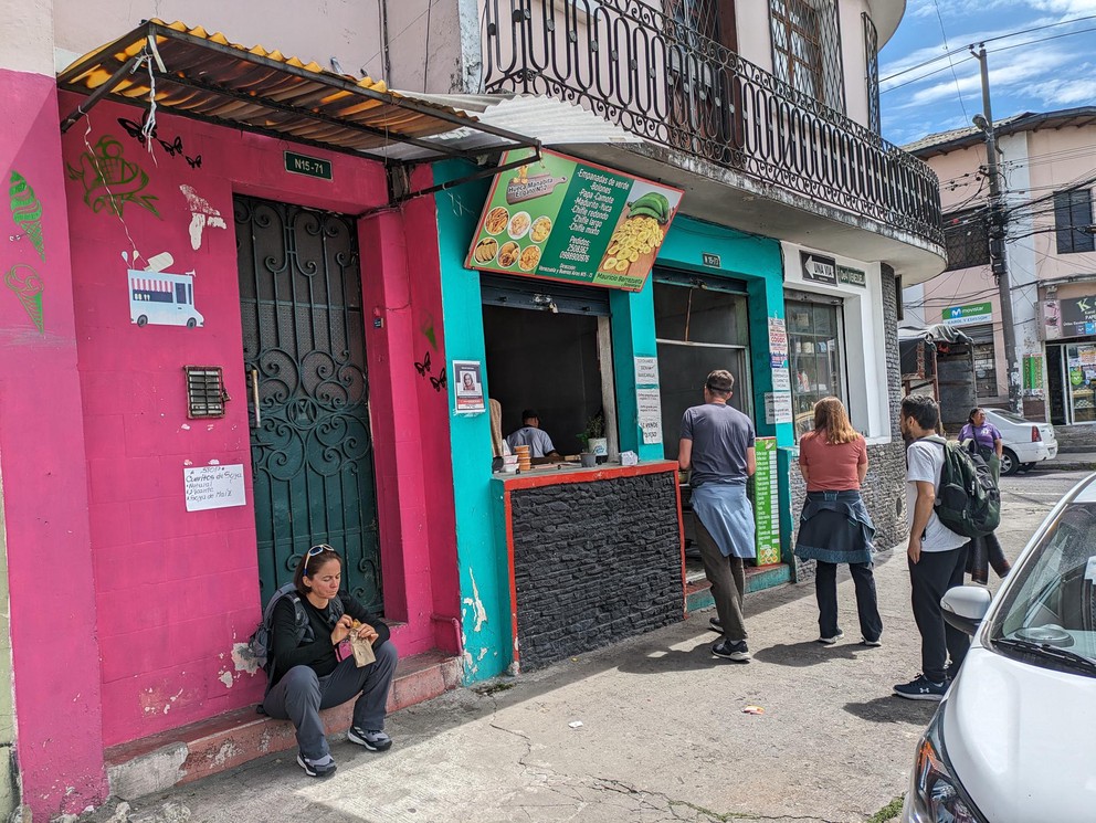 people wait on the sidewalk in front of a tienda selling empanadas
