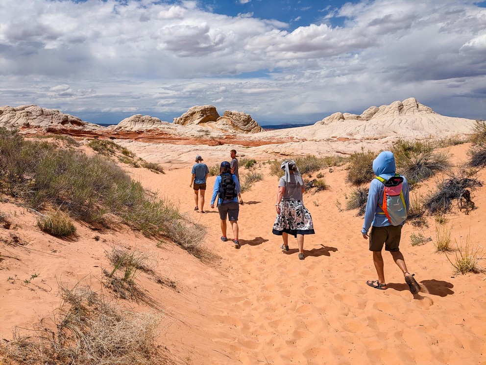 five hikers follow a sandy trail towards some white and red sandstone outcroppings